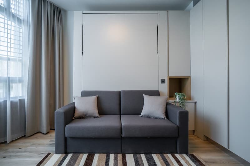 Spacious living room with large windows, gray sofa, decorative pillows, and a built-in storage unit with a plant