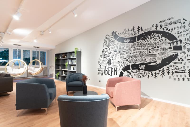 Spacious living room with large windows, hanging chairs, gray and pink armchairs, wooden shelves, and a large mural wall with illustrations of the city of Bern and surrounding areas.