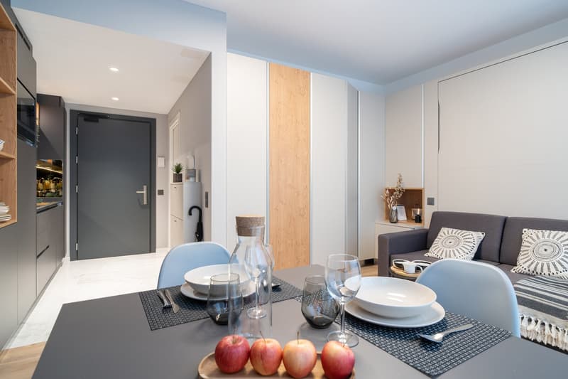 The image shows a modern and stylish open-plan living and dining area. The space features a gray sofa, a dining table with place settings, and a kitchen area with a dishwasher visible. The walls are a mix of white and wood paneling, creating a warm and in