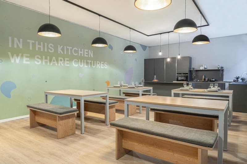 Modern kitchen with wooden tables and benches, black pendant lights, and a wall mural that says 'In this kitchen we share cultures'