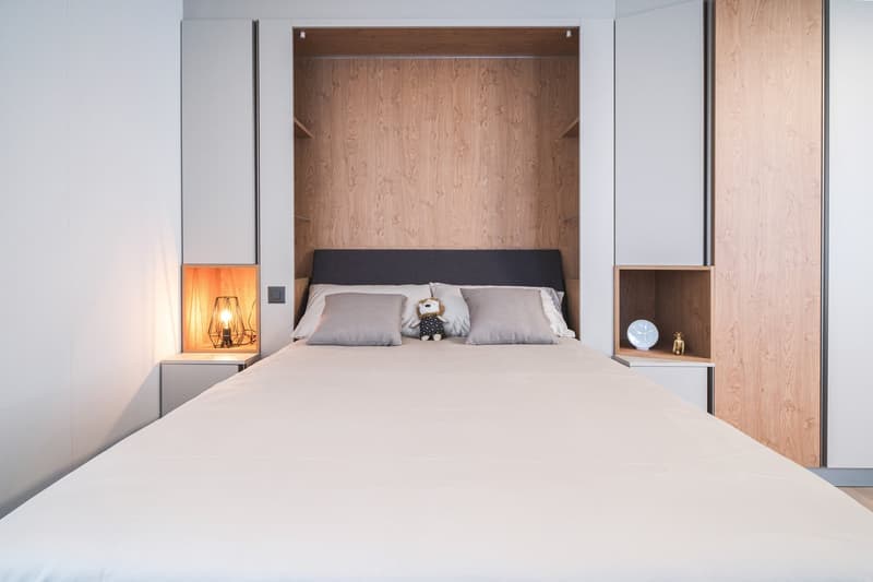 Spacious bedroom with a built-in wooden wardrobe, a large bed with gray bedding, and a decorative lamp on the nightstand. The room has a minimalist and modern design with a cozy atmosphere.