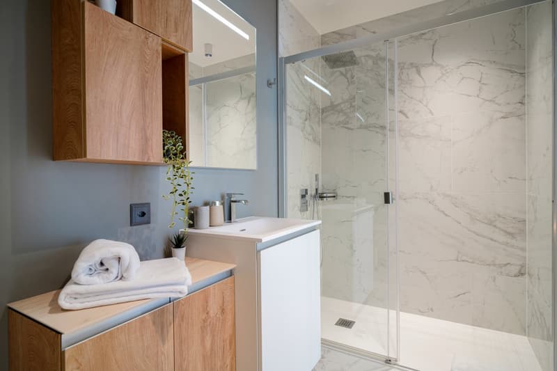 Modern bathroom with marble-like tile walls, a glass shower enclosure, a white vanity with a sink, and wooden storage cabinets. There are towels and a potted plant visible as well.