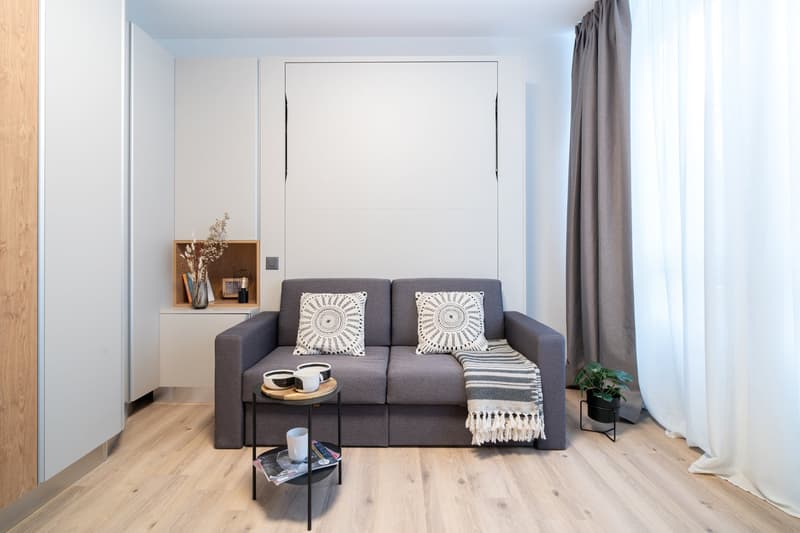 Gray sofa with patterned throw pillows, wooden shelving unit, coffee table, potted plant, and large window with curtains