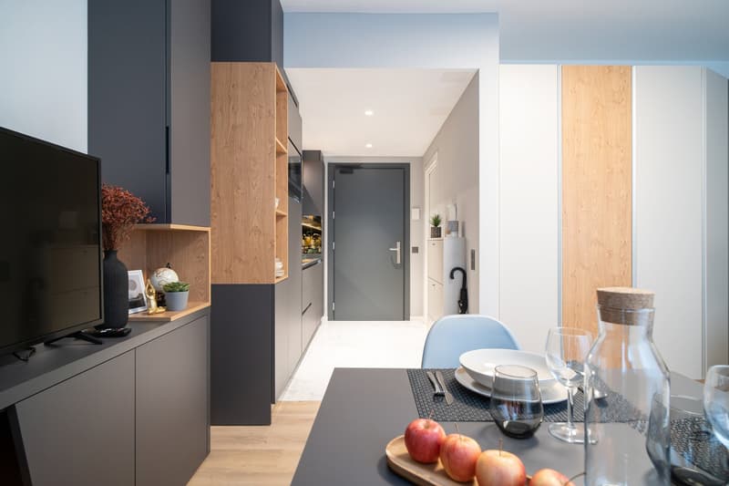 Modern kitchen with dark gray cabinets, wooden shelves, and a dishwasher. The kitchen opens up to a living area with a TV stand and a dining table with chairs.