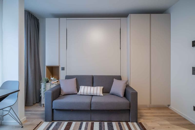Compact studio apartment with a gray sofa, built-in storage cabinets, and a large window with curtains providing natural light. The room has a minimalist and modern design aesthetic.