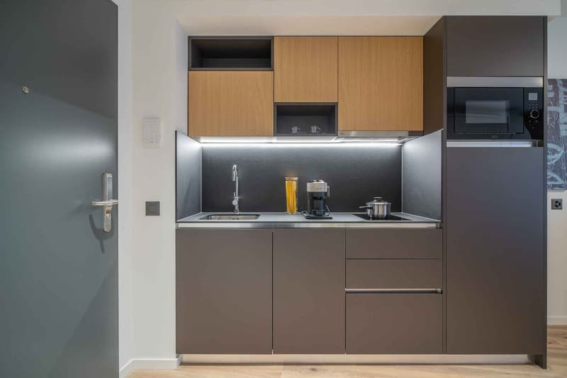 Compact kitchen with modern, minimalist design. Features wood-tone upper cabinets, gray lower cabinets, stainless steel appliances including a dishwasher, and a sleek countertop with a sink and cooking utensils.