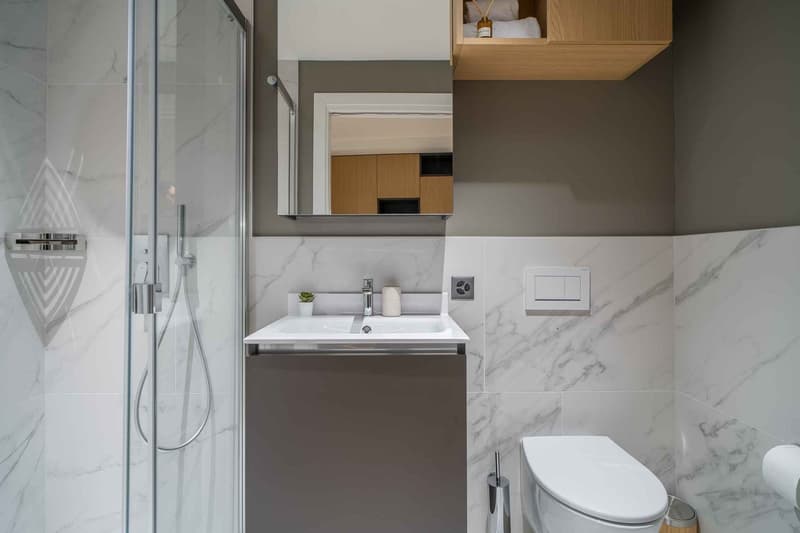 Modern bathroom with marble-like tiles, glass shower enclosure, vanity with sink, and built-in storage cabinet above the sink