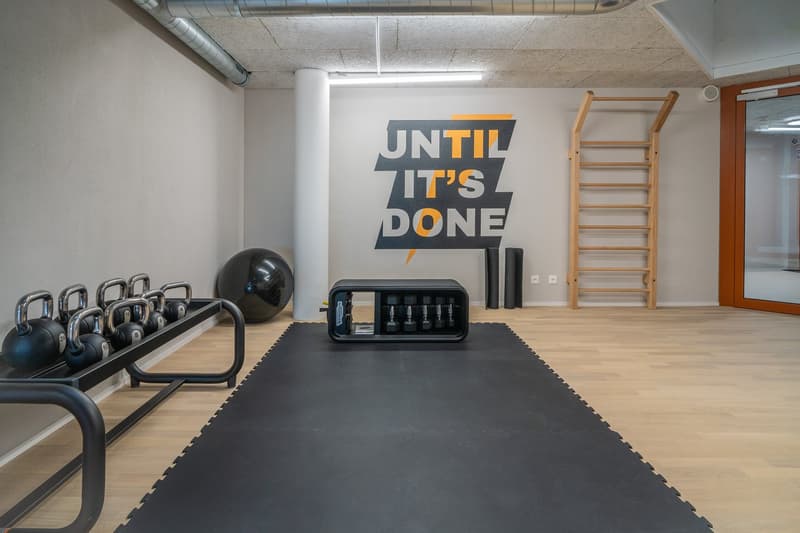 The image shows a gym or fitness room with various exercise equipment such as kettlebells, a weight rack, and a large motivational wall sign that says 'Until it's done'. The room has a concrete ceiling, wooden flooring, and a large window. The space appea