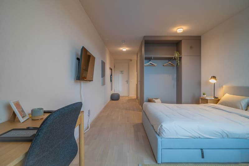 Spacious bedroom with hardwood flooring, white walls, large windows, a bed with blue bedding, a desk with a computer, and a closet with hanging space.