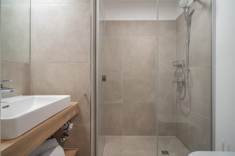 Modern bathroom with beige tile walls, a white vessel sink on a wooden vanity, and a glass-enclosed shower with a rainfall showerhead.