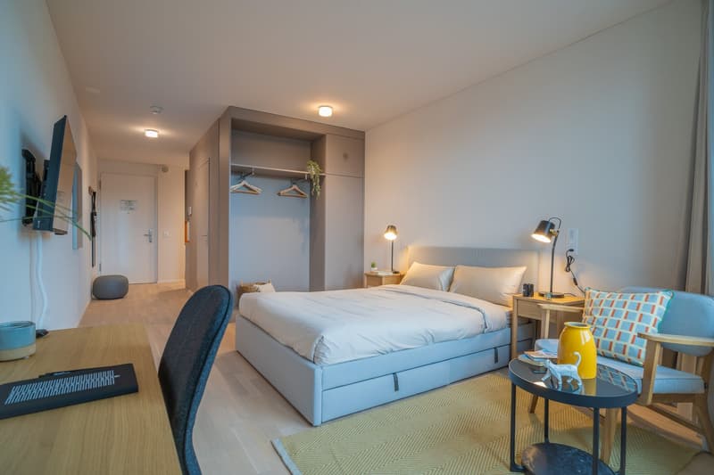 Spacious bedroom with a double bed, nightstands, lamps, and a TV mounted on the wall. There is a wardrobe closet and a seating area with a chair and small table. The room has large windows and a neutral color scheme.