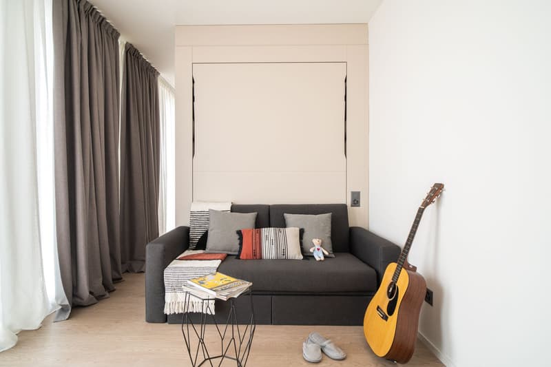 Gray sofa with decorative pillows, acoustic guitar, books on a side table, large windows with gray curtains