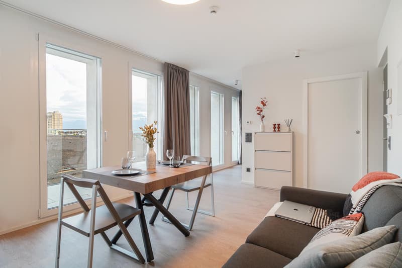 Spacious living room with large windows, wooden dining table, sofa, and storage cabinet. The room has a modern and minimalist design with neutral colors.