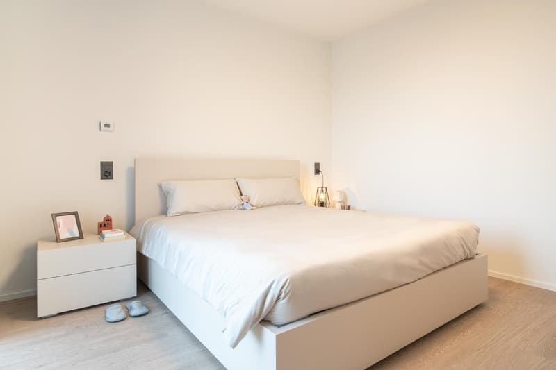 Spacious bedroom with a large bed, white bedding, and a nightstand. The room has a minimalist and clean design with white walls and hardwood flooring.