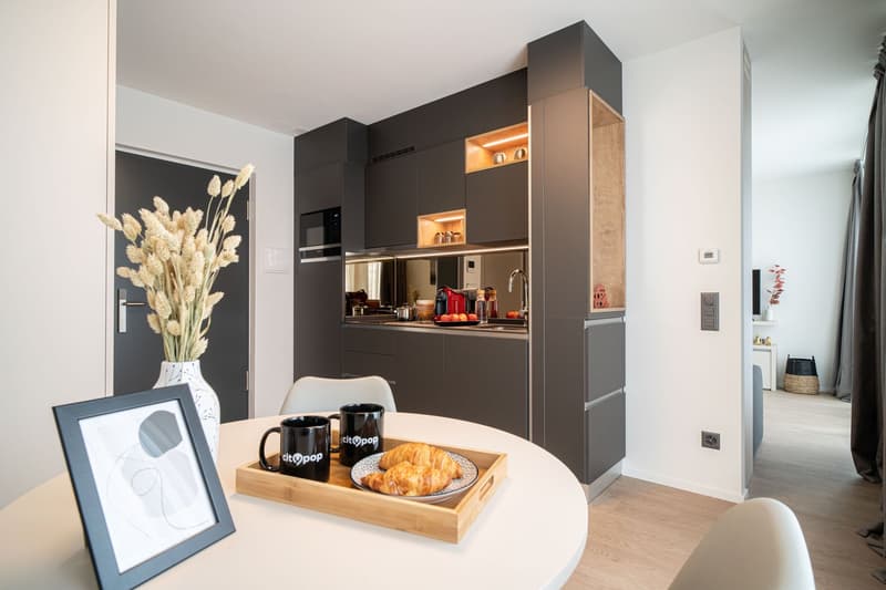 Modern kitchen with dark cabinets, built-in appliances, and a dining area with a round table and chairs. The kitchen features a dishwasher and is decorated with a vase of dried flowers and framed artwork on the wall.