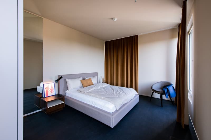 The image shows a modern and minimalist bedroom with a gray upholstered bed, white bedding, and brown curtains covering the window. There is a small side table with a lamp and a framed artwork on the wall. The room has a clean and simple design with a dar