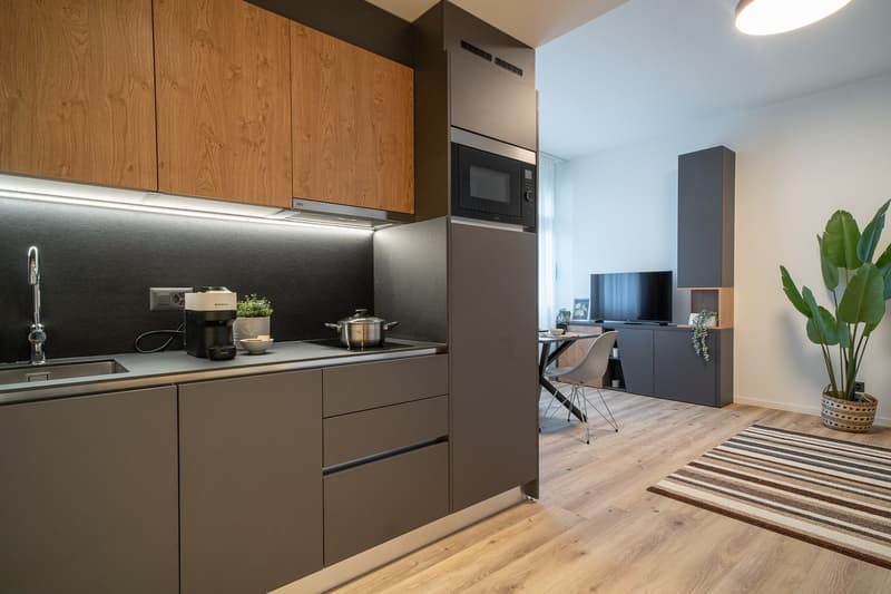 Modern kitchen with wooden cabinets, gray drawers, built-in microwave, and appliances on the countertop. The kitchen opens up to a living room area with a TV and furniture.