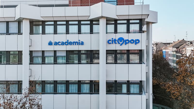 A modern, multi-story office building with large windows and signage for 'Macademia' and 'Citypop' companies. The building has a clean, contemporary architectural style with a mix of glass and white panels.
