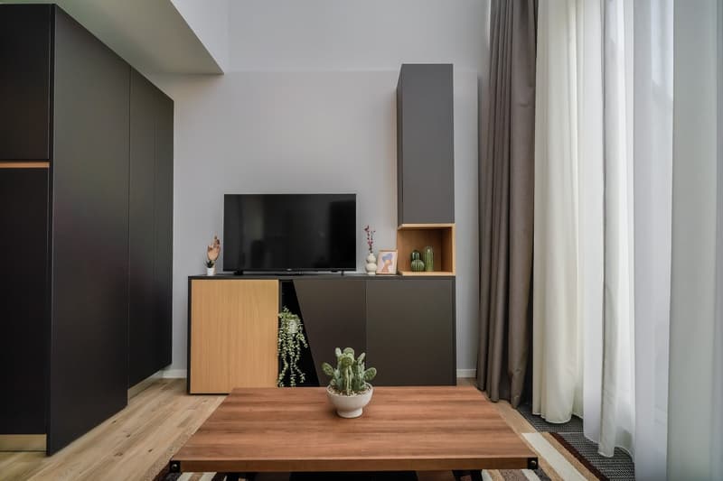 Modern living room with a large TV unit, wooden table, and potted plants. The room features dark-colored cabinets, white curtains, and a fireplace.