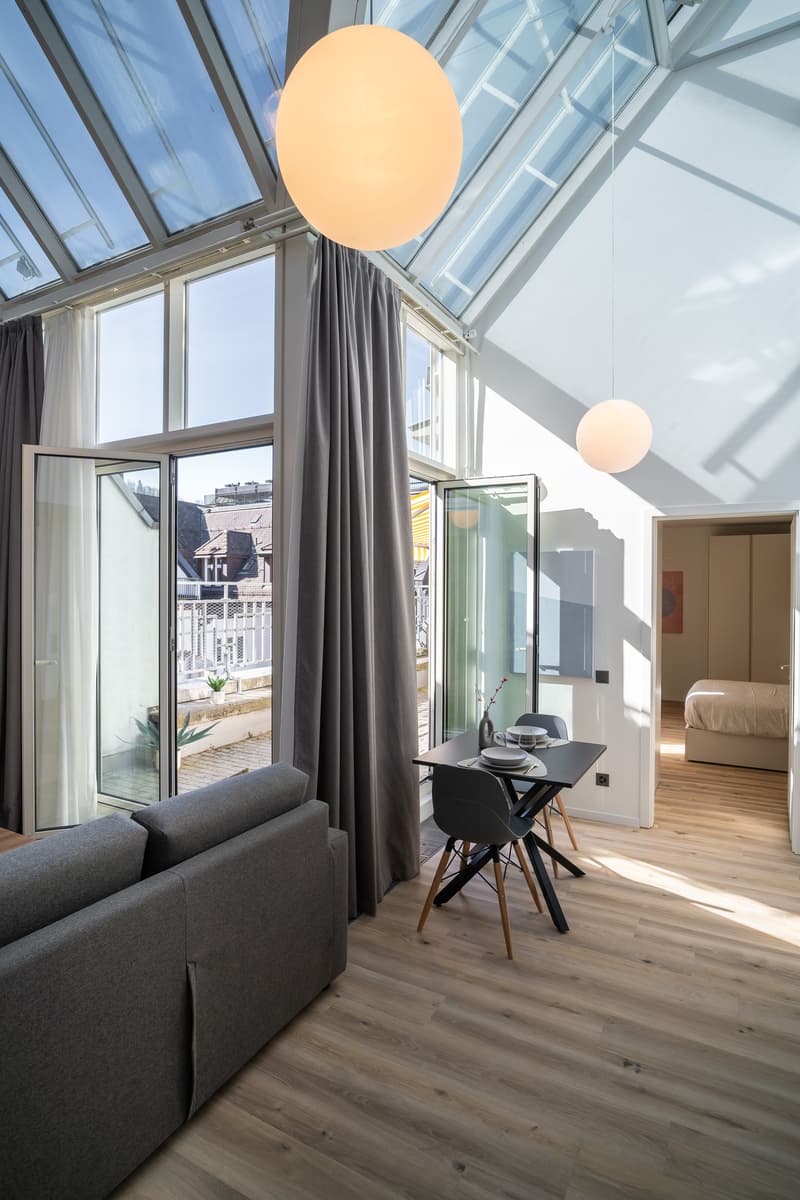 Spacious living room with large windows, glass doors leading to a balcony, gray sofa, dining table with chairs, and pendant lighting fixtures.