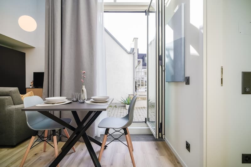 Modern dining area with a wooden table, black metal frame, and gray chairs. Large windows provide natural light and access to a balcony or terrace. The room also features a sofa and TV in the background.