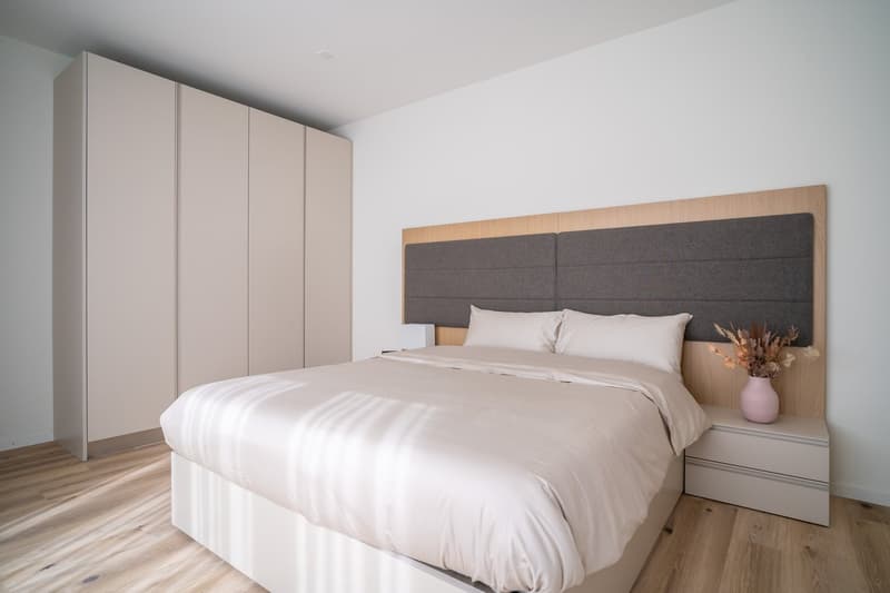 Spacious bedroom with a large bed, gray upholstered headboard, and built-in wardrobe. The room has hardwood floors and a neutral color scheme with white bedding and accents of wood and gray. There appears to be a balcony or terrace visible through the win