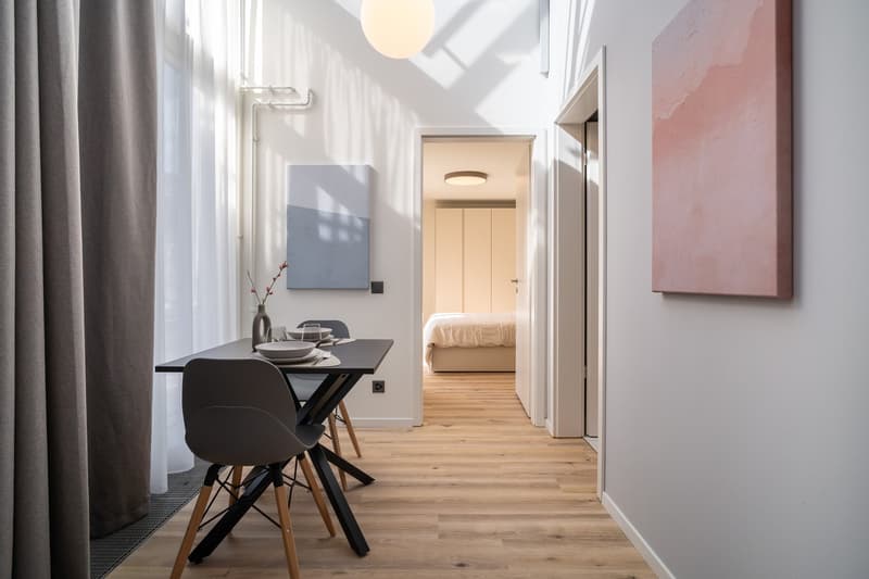 Open-concept living space with a dining area, kitchen, and bedroom visible. The apartment features hardwood floors, white walls, large windows, and modern furnishings including a black dining table and chairs, artwork on the walls, and a bed in the bedroo