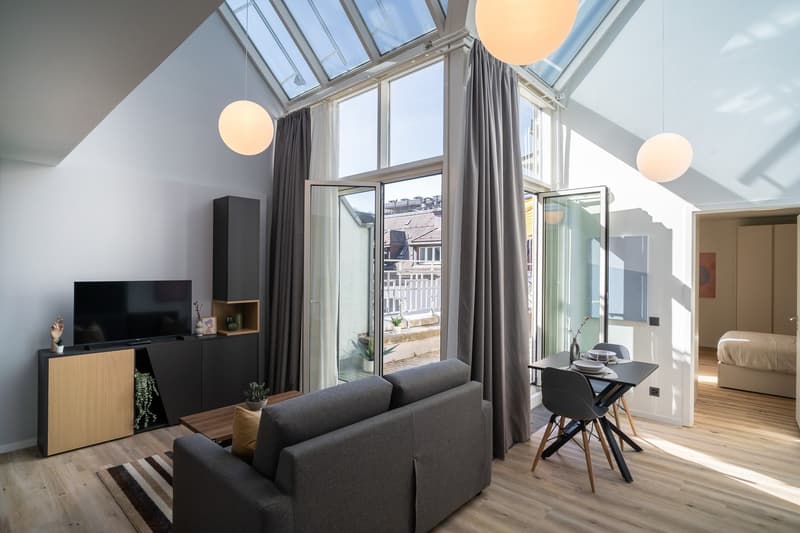 Spacious living room with high ceiling and large windows, providing ample natural light. The room features a gray sofa, a TV stand, and a dining table with chairs. The room also has a balcony or terrace accessible through the glass doors.