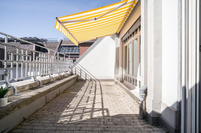 A covered balcony with a yellow and white striped awning, brick walls, and a paved walkway leading to the entrance.
