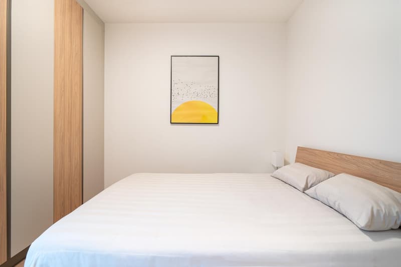 Double bed, bedside lamps, beige pillows, white bedspread, white walls, wooden nightstand, picture frame on wall