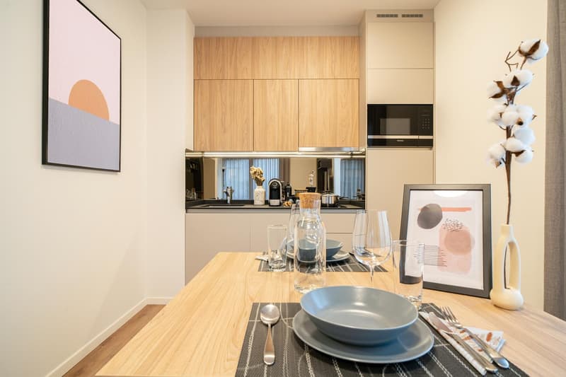 A modern dining area featuring a wooden table set for a meal. There are clear wine glasses, plates, cutlery, and a framed picture. Behind it, there is a kitchen with wooden cabinets, a black microwave, and a sink. A painting is on the wall.