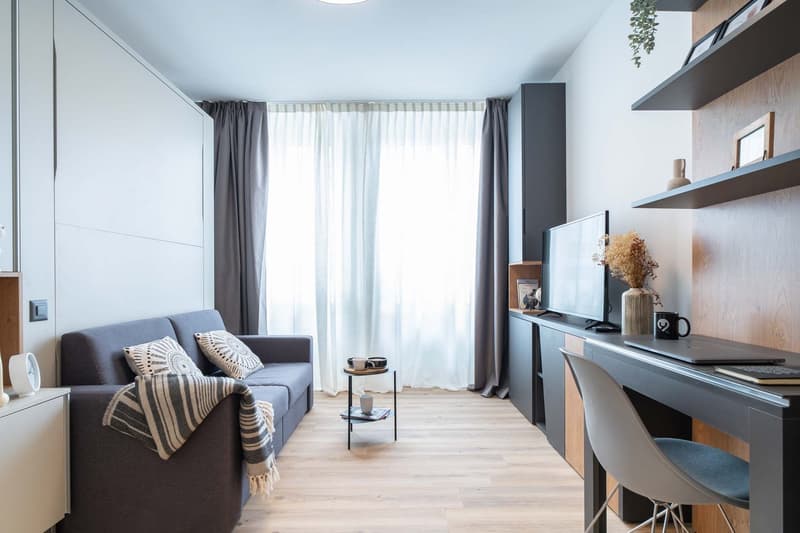 cozy grey sofa, large window with curtains, wooden floor, built-in entertainment unit, desk, and chair, floor lamp, open shelving with decor