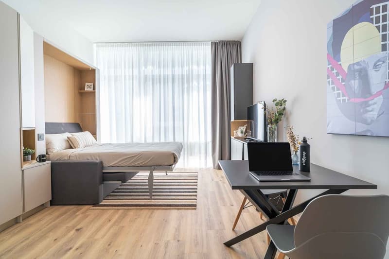 Bedroom with bed, sofa bed, desk, and chair, wooden floor, large window with curtains, and wall art.