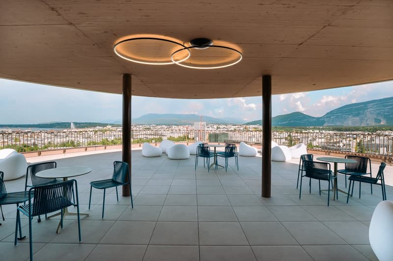 Open outdoor area with tables and chairs, circular lights, tiled floor, balcony with metal railing, panoramic city view