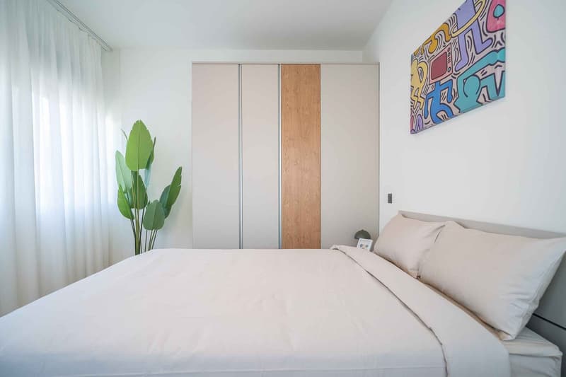 Bedroom with a bed, white walls, white curtains, a plant, a wardrobe, a picture frame, and a colorful painting on the wall.
