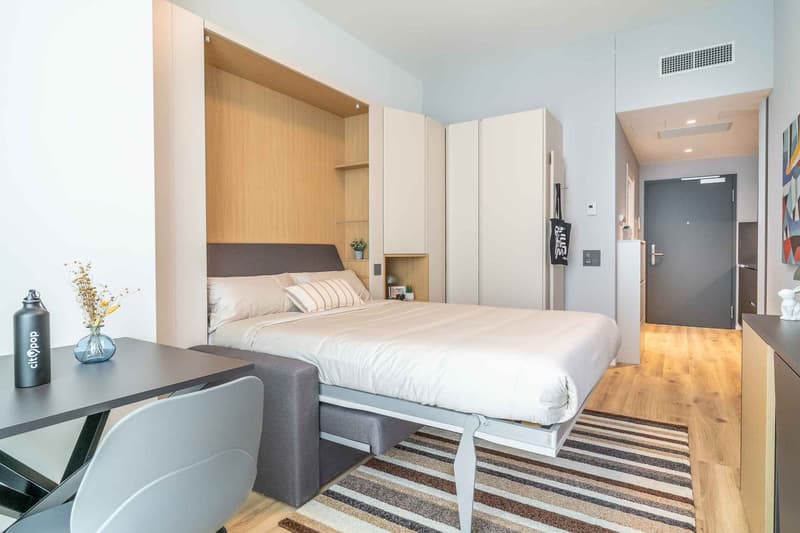 A bedroom with a bed, desk, chair, and a rug. The bed is made with white sheets and pillows. The desk has a vase with flowers and a water bottle. The room has wooden floors and white walls. There is a closet with shelves and a door leading to another room