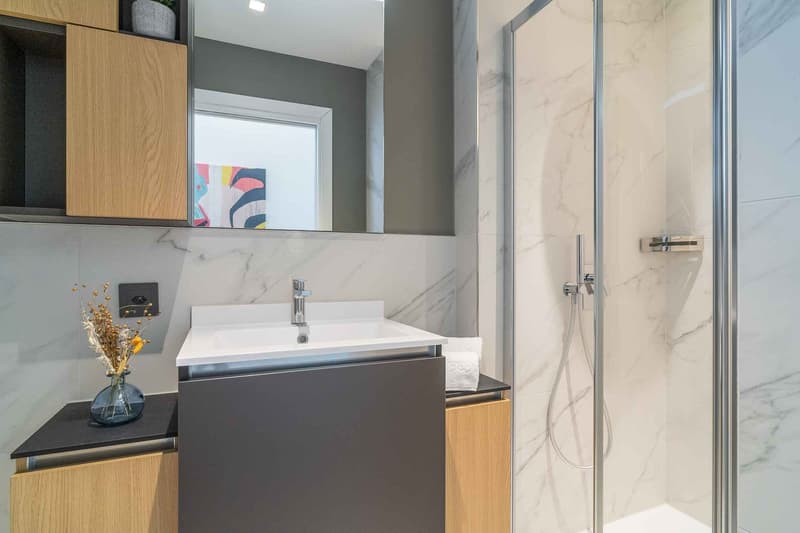 modern bathroom with marble tiles, white sink, shower, wooden cabinets, power outlet, vase with flowers