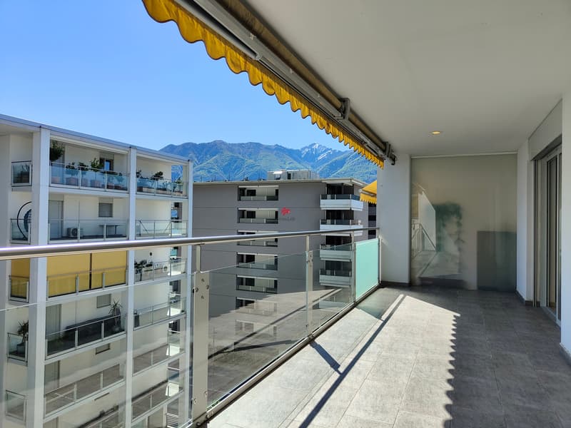 Der Blick von der Terrasse schweift über mehrere Berggipfel, und zwischen den Gebäuden ist ein kleiner Teil des Sees zu erkennen / La vista dalla terrazza spazia su diverse cime delle montagne e un