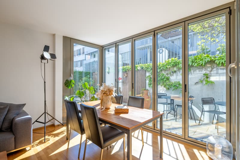 Ein helles Wohnzimmer mit bodentiefer Vollverglasung bietet einen offenen Blick auf einen gemütlichen Sitzplatz und schafft....