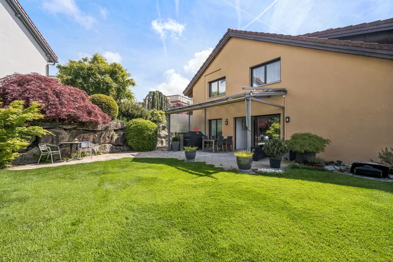 Die gedeckte Gartenterrasse lädt zum Entspannen, Geniessen und Verweilen ein. / the backyard of a home with a large green lawn in the front yard