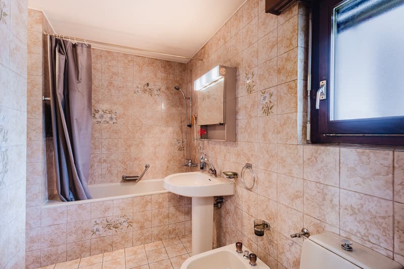 Das Badezimmer in L-Form verfügt über eine Waschküchenecke, die durch eine Wand von der Badewanne abgetrennt ist. / Il bagno a forma di L ha un angolo lavanderia separato dalla vasca da bagno da un