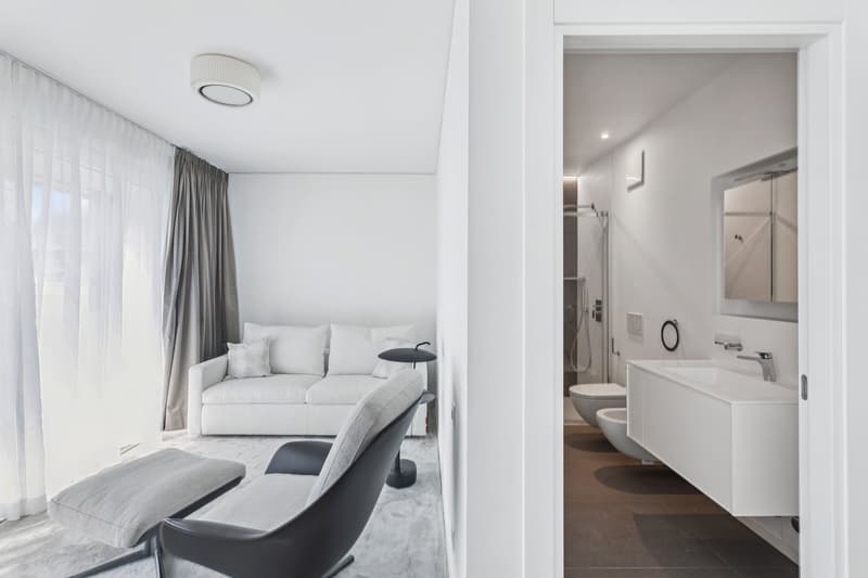 modern living room with white couch, black chair, white ottoman, white walls, white curtains, white bathroom with sink, toilet, and shower