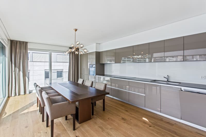 Dining room with wooden floor, large windows, dining table, chairs, chandelier, modern kitchen with gray cabinets, sink, dishwasher, and oven.
