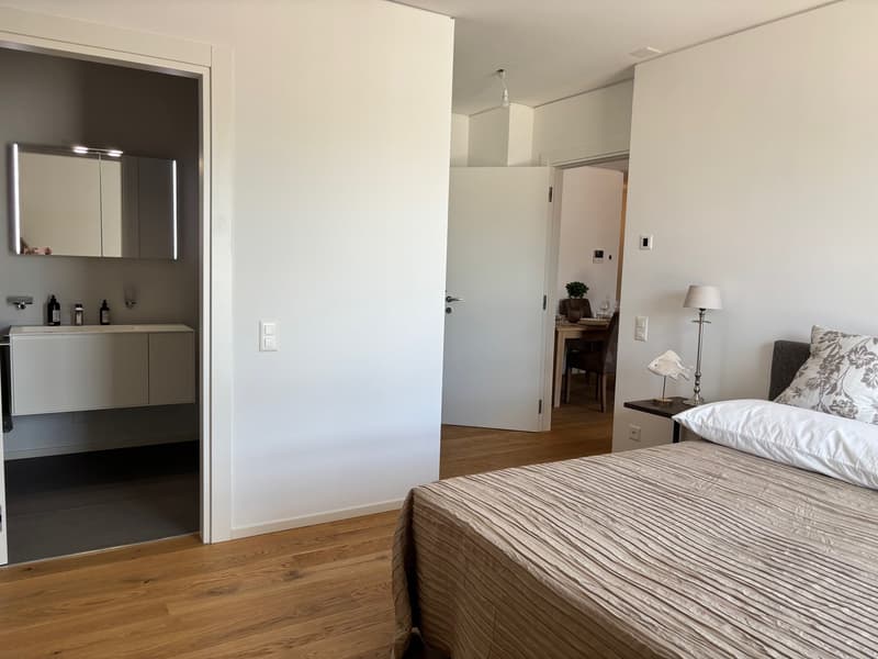 Spacious bedroom with hardwood floors, white walls, large bed with striped bedding, bedside table with lamp, and a bathroom visible through the open doorway.