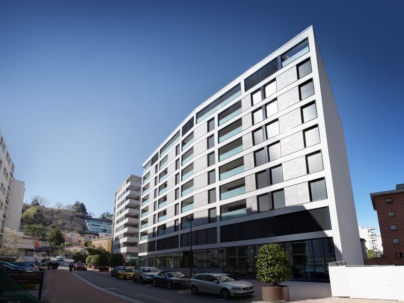 Modern multi-story building with a curved facade, large windows, and a ground floor with commercial spaces. There is a parking area in front of the building.
