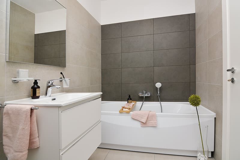 modern bathroom with bathtub, gray tiled walls, vanity with sink, mirror and towels