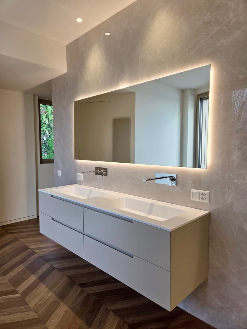 modern double sink vanity, large mirror with lighting, sleek faucets, white cabinetry, marble-like wall finish, power outlet, wooden flooring