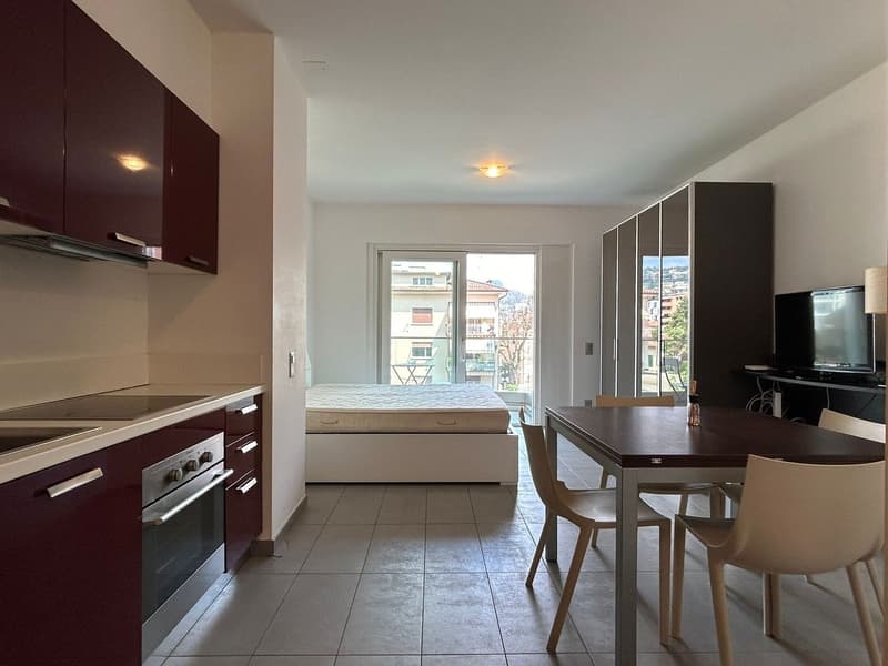 Kitchen with dining area, compact bedroom with bed, sliding doors leading to a balcony, white tiles, modern appliances, bed storage unit