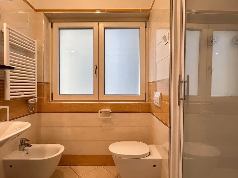 A modern bathroom with white tiles and fittings, featuring a window, sink, bidet, and toilet.