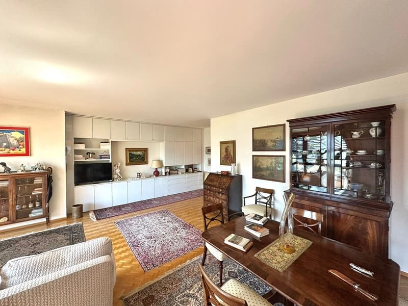large living room with wooden dining table, 2 sofas, wooden chairs, white cupboards, wooden desk, glass cabinet, decorative items, framed pictures, carpet